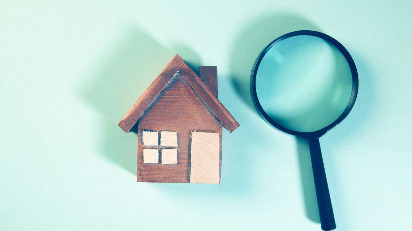 Modellino di casetta di legno accanto a una lente di ingrandimento su fondo azzurro